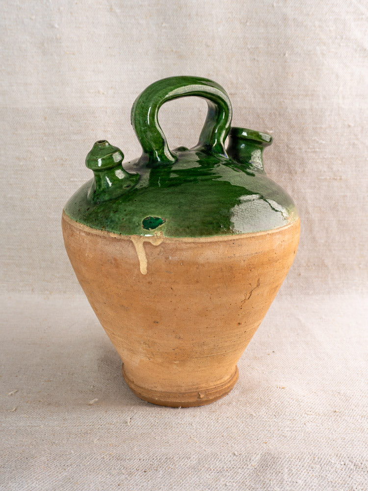 Antique French gargoulette with emerald green glaze, twin spouts, curved handle, unglazed clay base on textured fabric.