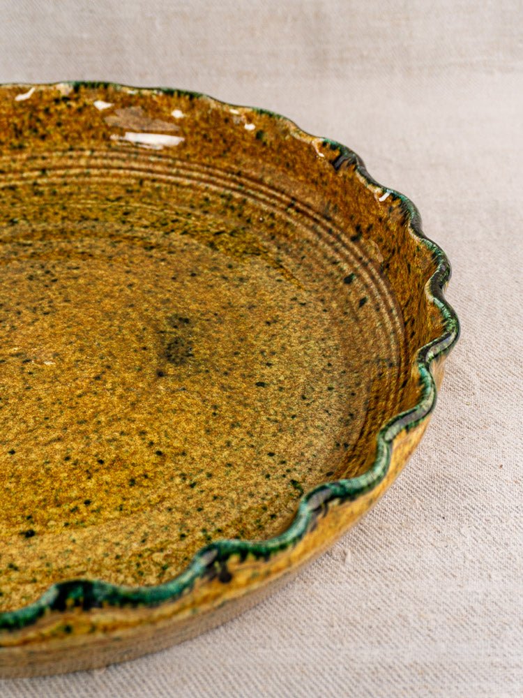 Vintage French JANY Studio earthenware tarte dish with scalloped edge, brown-green glaze on beige fabric.
