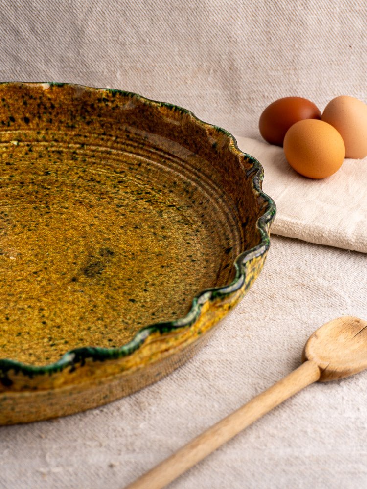 French JANY Studio earthenware tarte dish beside wooden spoon and brown eggs on beige cloth.