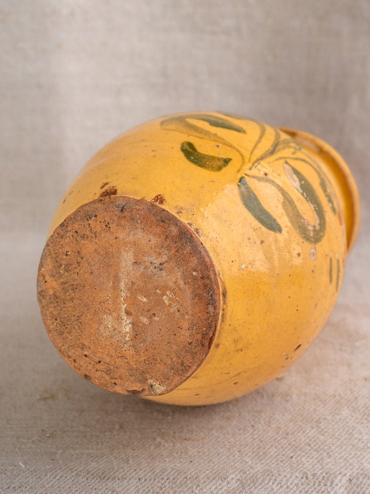 Antique French yellow glazed pitcher with hand-painted floral earthenware, showing unglazed base on beige fabric.