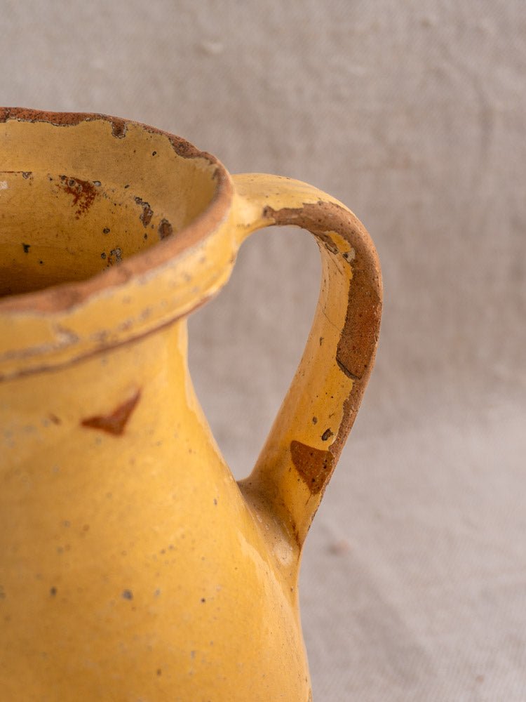 Antique French yellow glazed ceramic pitcher with hand painted floral design, chips, and patina on beige fabric.