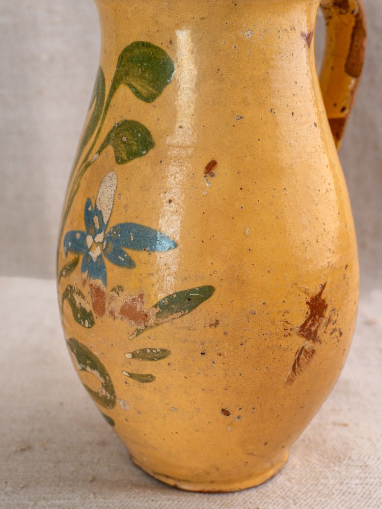 Antique French yellow-glazed earthenware pitcher with blue floral design, minor chips, on fabric-textured background.