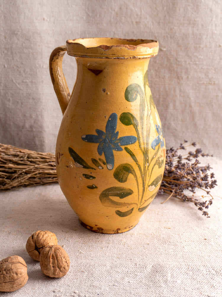 French yellow glazed ceramic pitcher hand painted with blue flowers and green leaves on beige fabric.