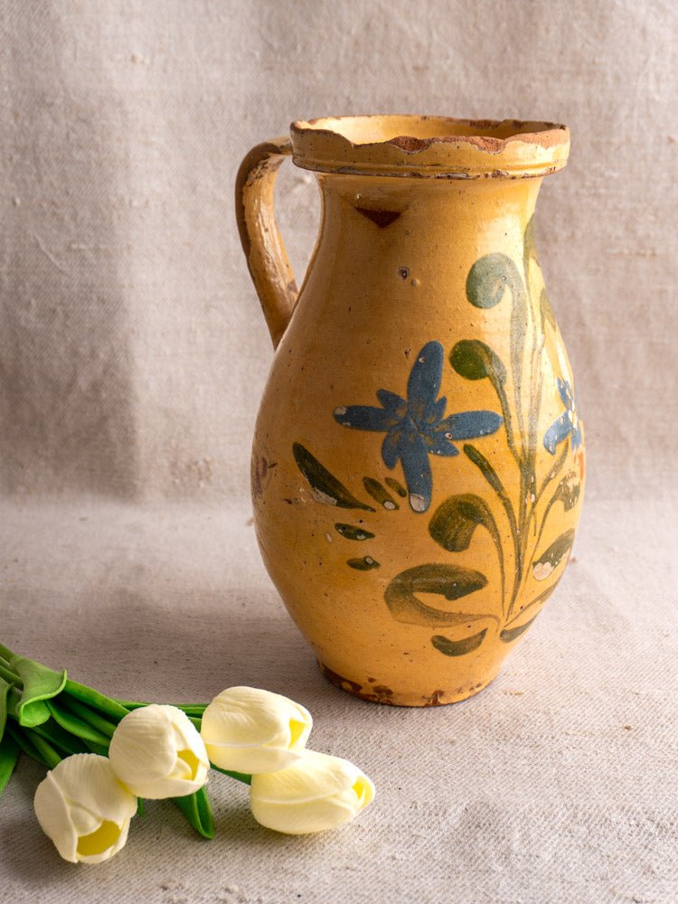 Antique French yellow glazed earthenware pitcher with hand-painted blue and green floral motifs, beside white tulips.