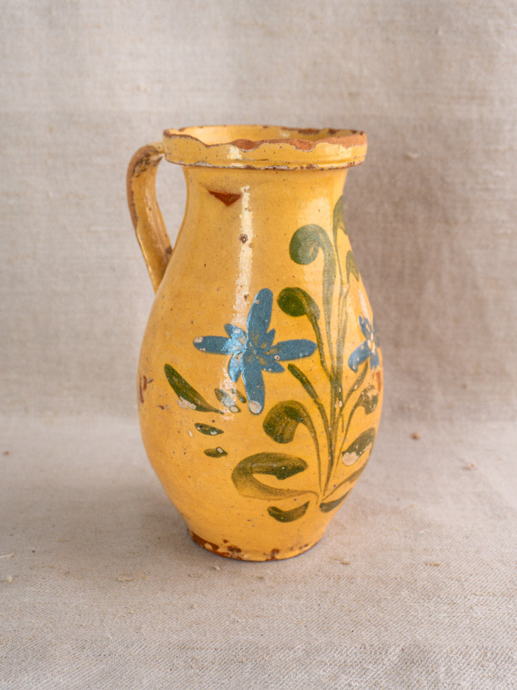 French yellow glazed ceramic pitcher hand painted with blue flowers and green leaves on beige fabric.