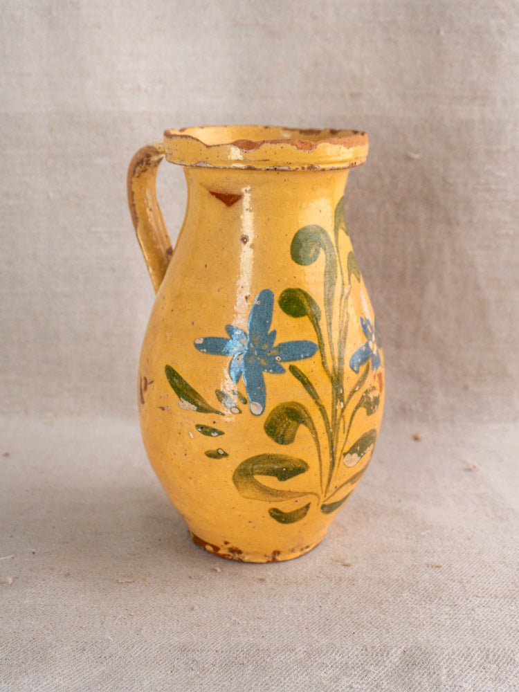 French yellow glazed ceramic pitcher hand painted with blue flowers and green leaves on beige fabric.