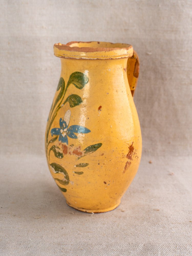 The Antique French Yellow Glazed Pitcher | Hand Painted Floral Earthenware, a small yellow ceramic piece featuring a handle and hand-painted with blue flowers and green leaves, sits on a neutral fabric background—an exquisite example of antique French pitcher craftsmanship.
