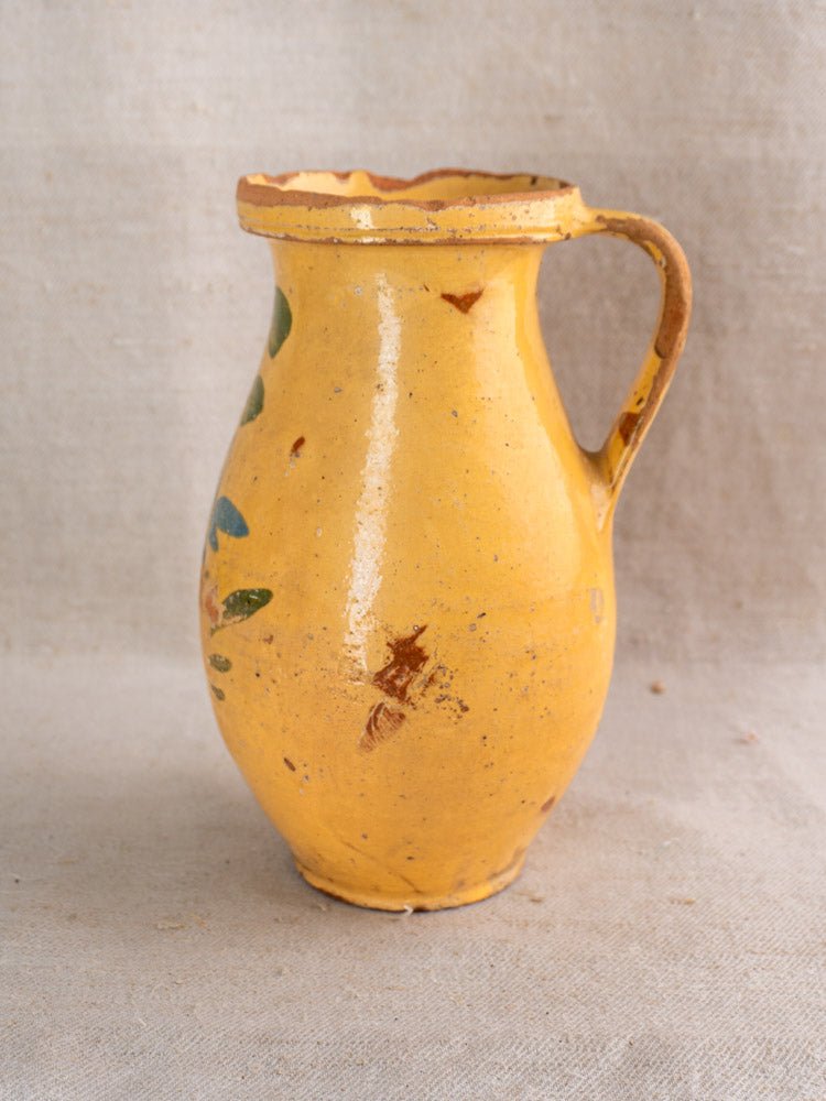 An Antique French Yellow Glazed Pitcher—crafted from earthenware with a handle and hand-painted floral motifs—sits on a neutral fabric background, its slightly worn finish embodying the charm of classic French country pottery.