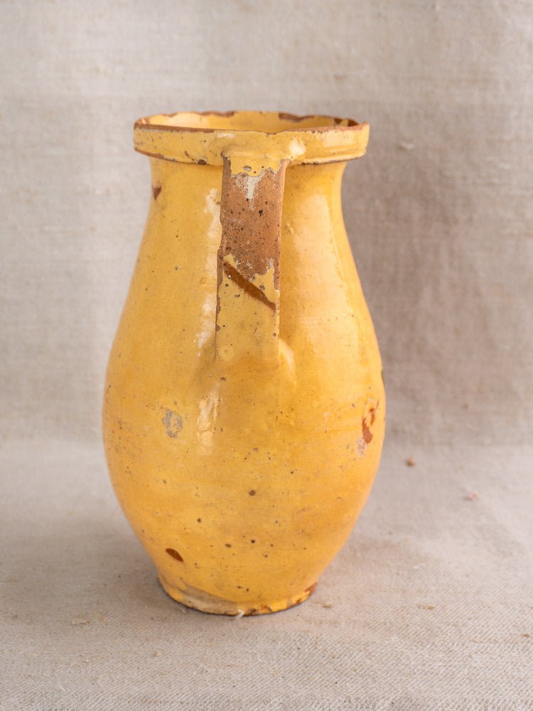 Antique French yellow glazed ceramic pitcher with floral accents and worn areas, on beige fabric.