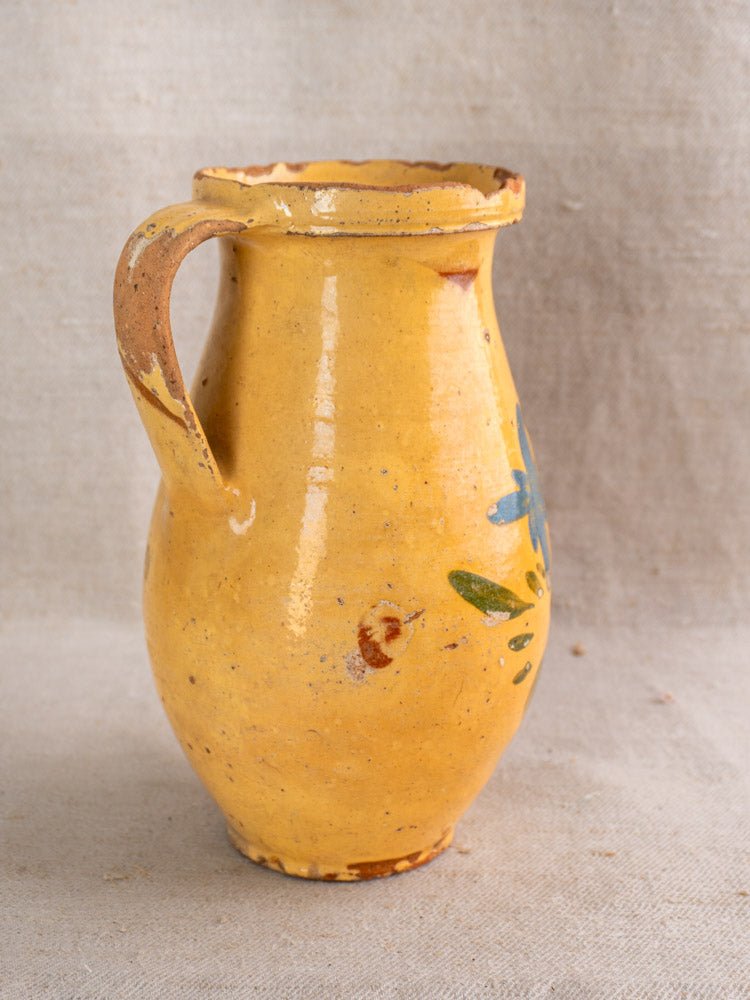 Antique French yellow glazed ceramic pitcher with hand-painted blue and green floral details on beige fabric.