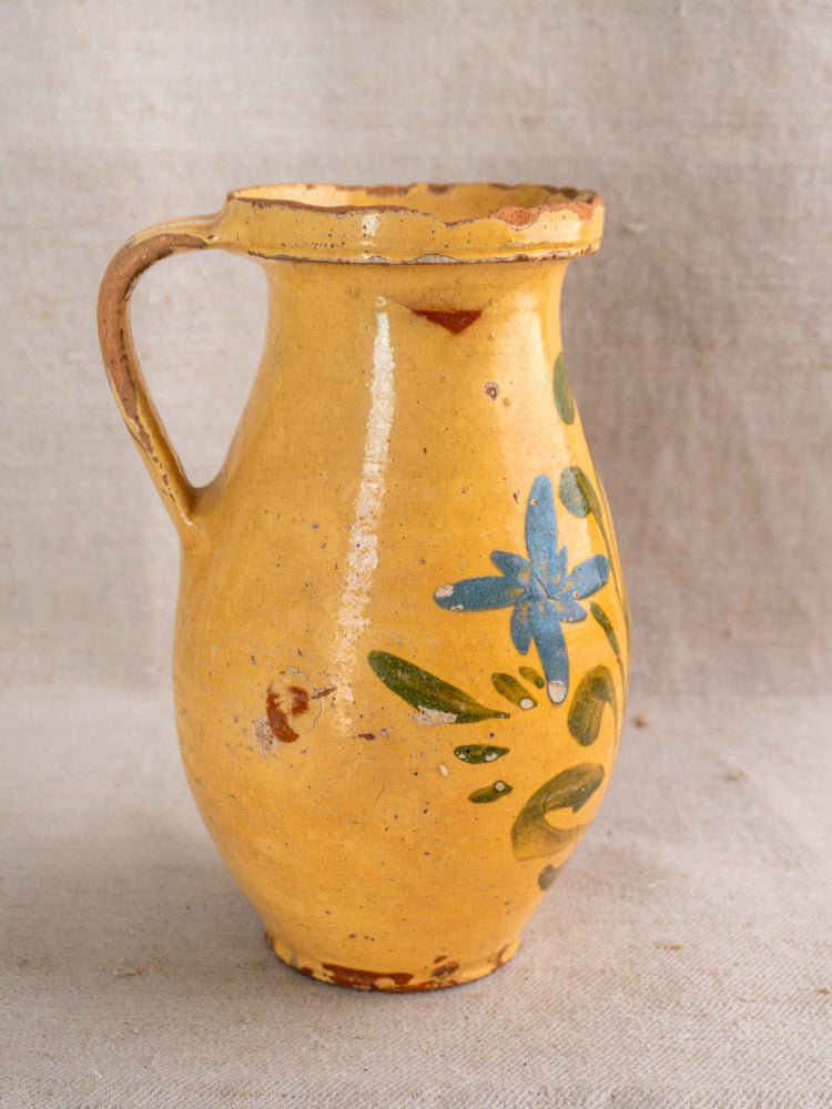 The Antique French Yellow Glazed Pitcher, a hand-painted earthenware piece featuring a handle, green leaves, and a blue flower motif, evokes classic French country pottery as it rests on a beige textured surface.