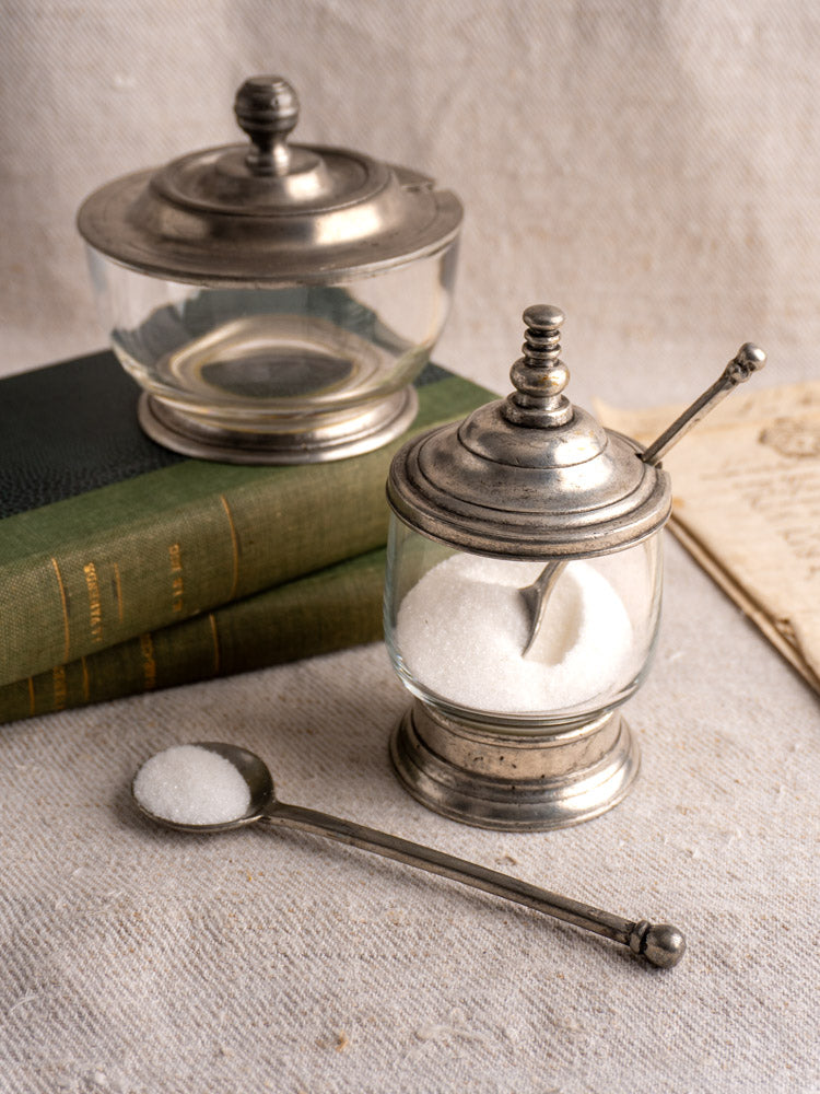 Pair of vintage French pewter and glass condiment jars with spoons, one on old green books, the other on textured fabric.