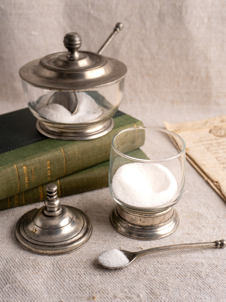 Vintage French pewter and glass condiment jars with spoons on beige fabric, green books and paper in background.