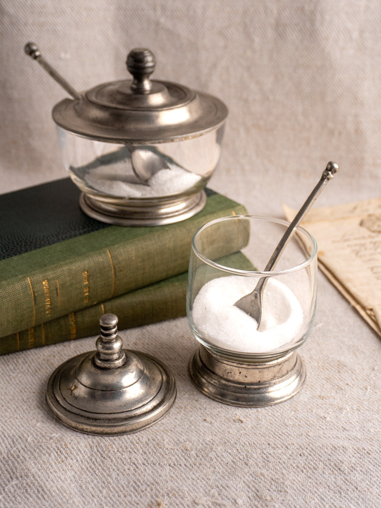 Pair of vintage French pewter and glass condiment jars with spoons on green books and linen.
