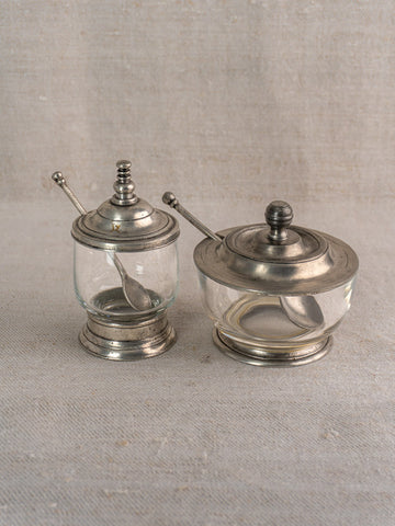 Pair of vintage French glass and pewter condiment jars with spoons, side by side on neutral fabric.