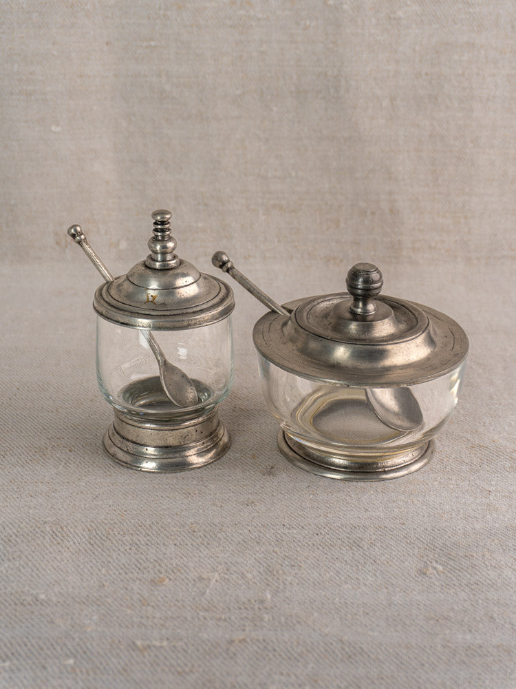 Pair of vintage French glass and pewter condiment jars with spoons, side by side on neutral fabric.