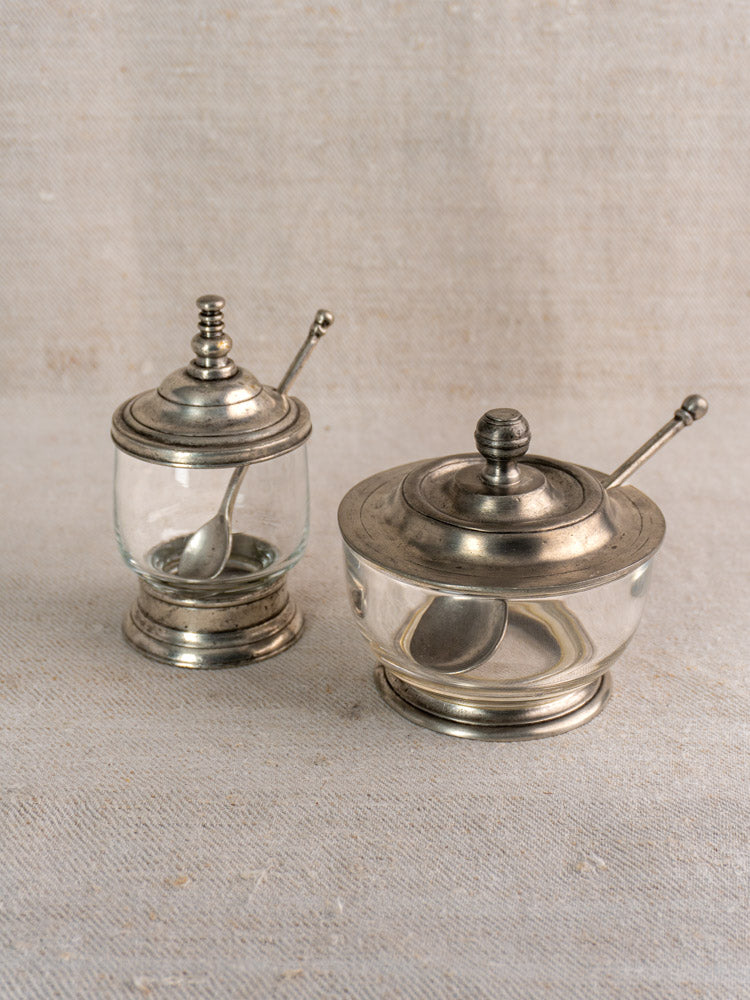 Pair of vintage French pewter and glass condiment jars with spoons, decorative finials, on beige fabric surface.