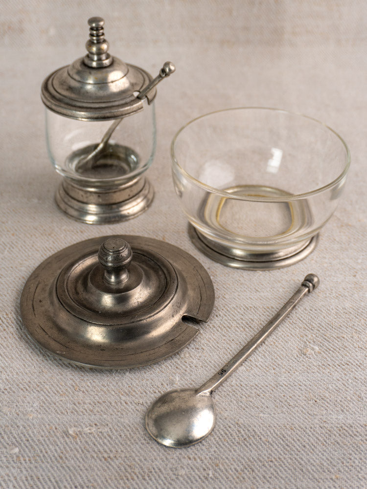 Set of two vintage French pewter and glass condiment jars with spoons on light textured fabric.