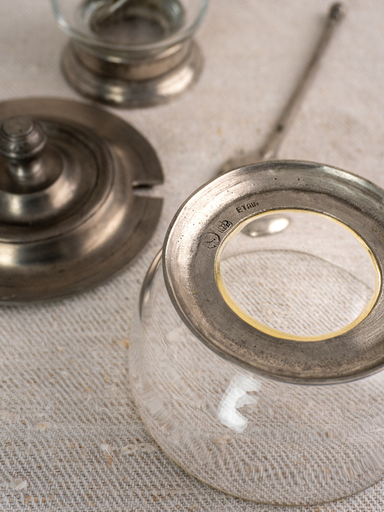 Pair of vintage French pewter and glass condiment jars with spoons, hallmarked “ETAIN,” on textured beige fabric.