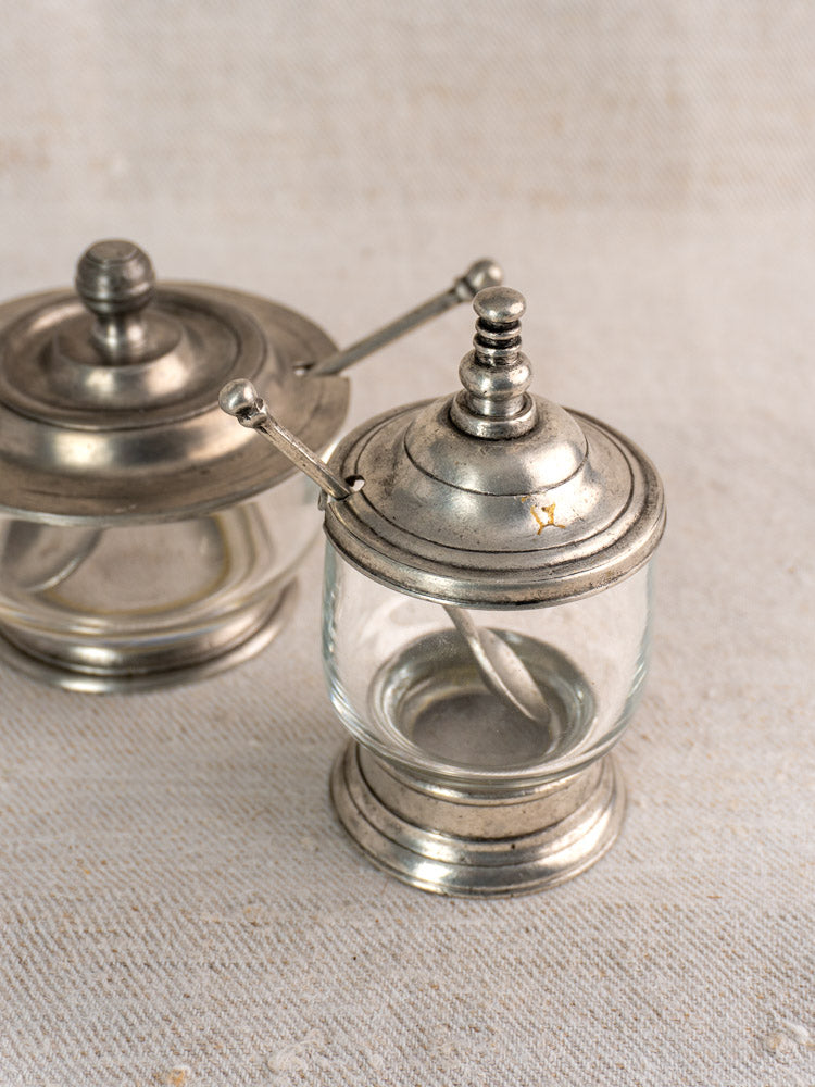 Vintage French pewter and glass condiment jars with spoons on textured beige fabric, one sharply focused, one softly blurred.