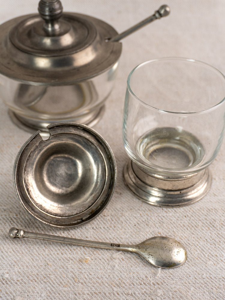 Pair of vintage French pewter and glass condiment jars with spoons on a light fabric surface.