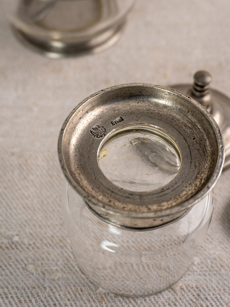 Vintage French pewter and glass condiment jar with spoon, from a tableware set, on a textured surface.