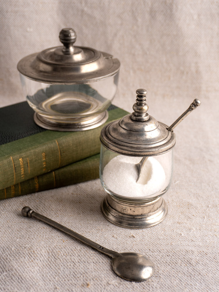 Pair of vintage French pewter and glass condiment jars with lids and spoons on green books.