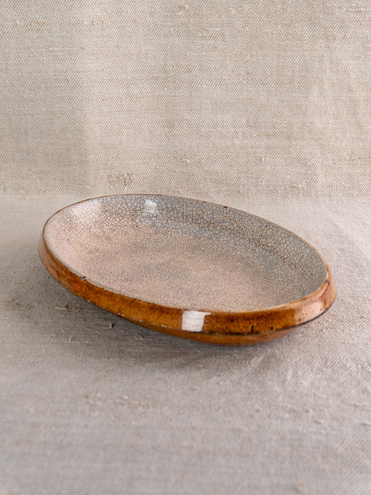 Antique French ceramic gratin dish with crackle-glaze beige interior, amber-brown exterior, on neutral linen fabric.