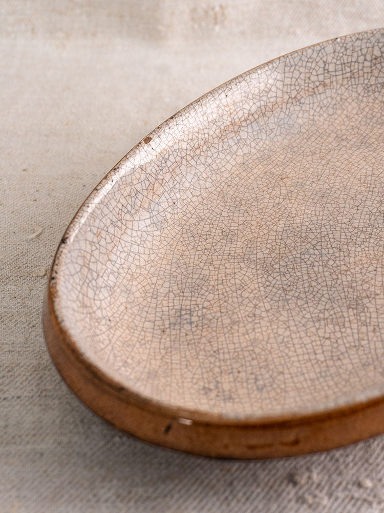 Antique French gratin dish with crackle glaze, earthy brown tones, and irregular form on beige textured fabric.