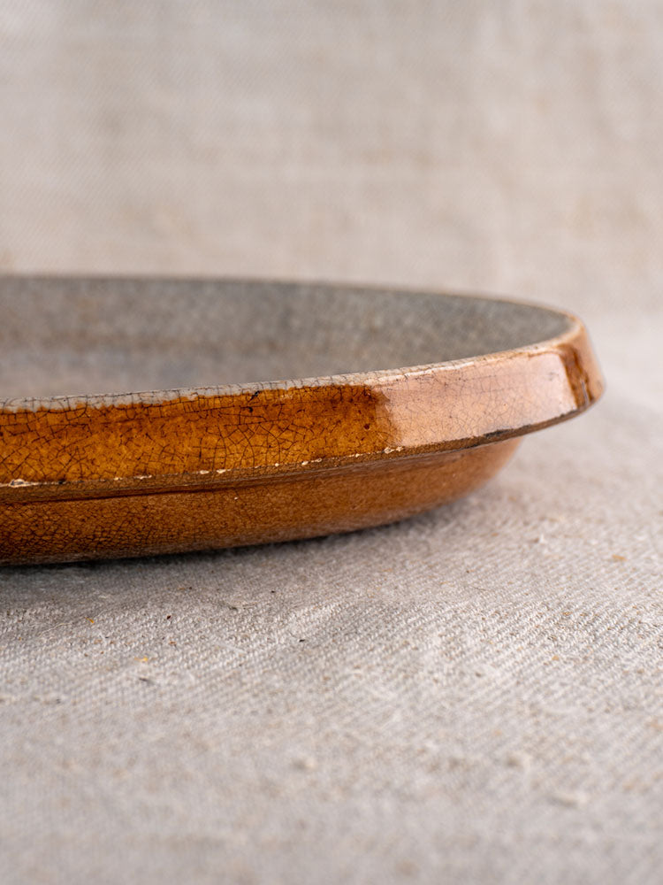 Antique French Provençal Tian gratin dish with brown crackle glaze rim on textured beige fabric background.