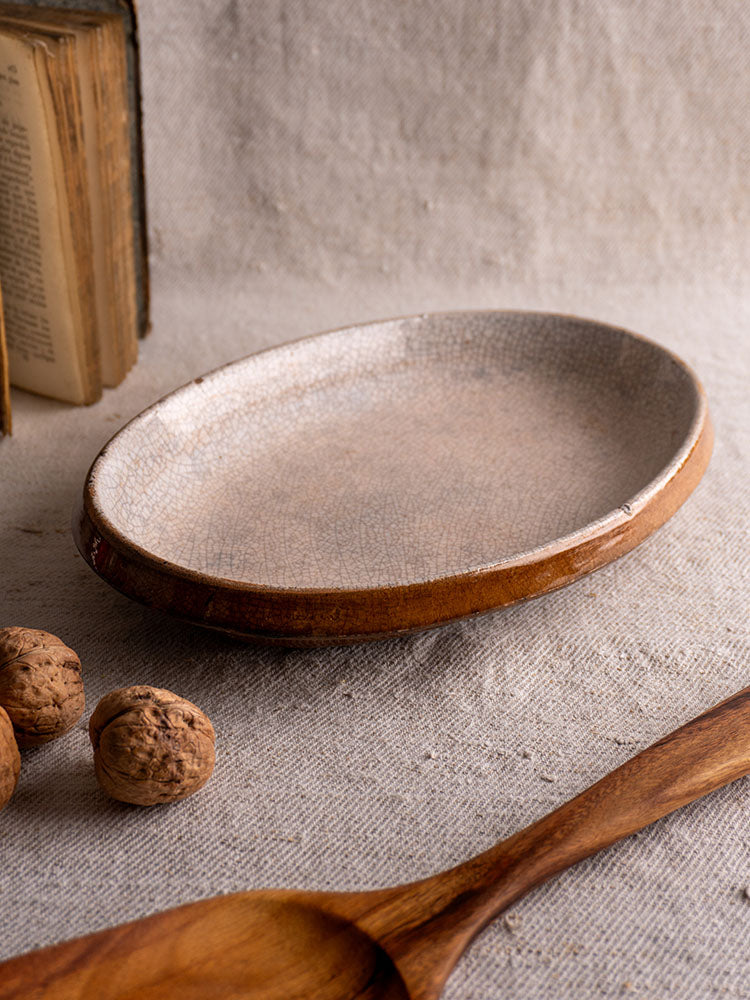 Antique French Provençal Tian with crackle glaze on textured fabric, wooden spoon, walnuts, and open book nearby.