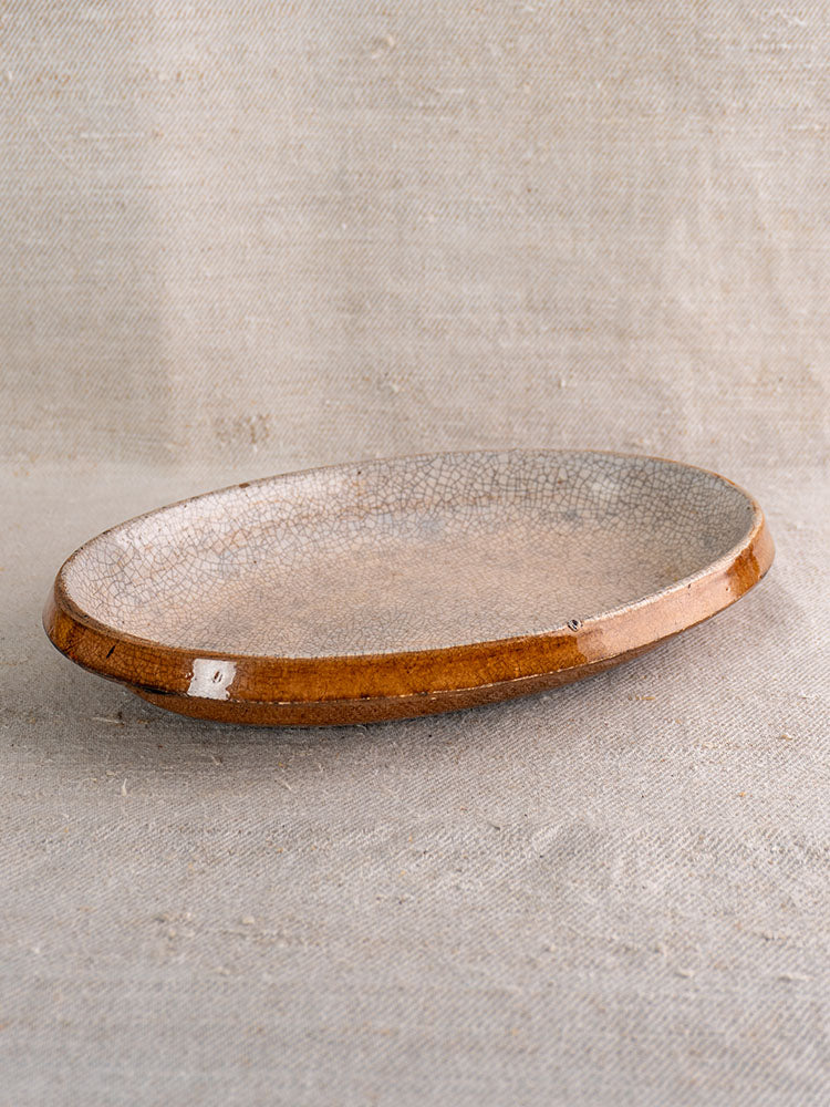 Antique French ceramic gratin dish with crackle glaze, earthy brown rim, white repair patch on textured beige fabric.