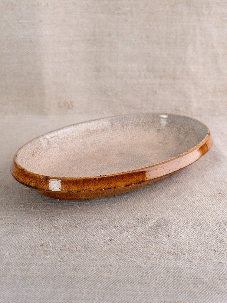 Antique French gratin dish with crackle glaze and brown rim, oval shape on textured beige fabric.