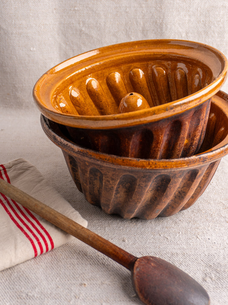 Antique French glazed Kouglof molds stacked, with wooden spoon and striped white cloth on beige fabric.