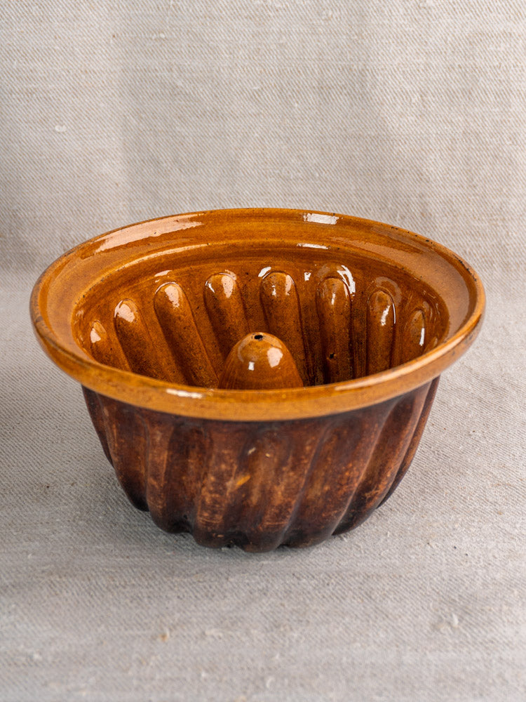 Antique French Kouglof mold with glossy brown glaze and fluted sides on fabric background.
