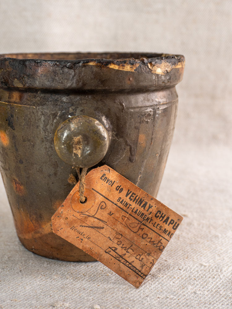 Antique French terracotta Pot à Graisse with original merchant tag and aged French script on beige textured fabric.