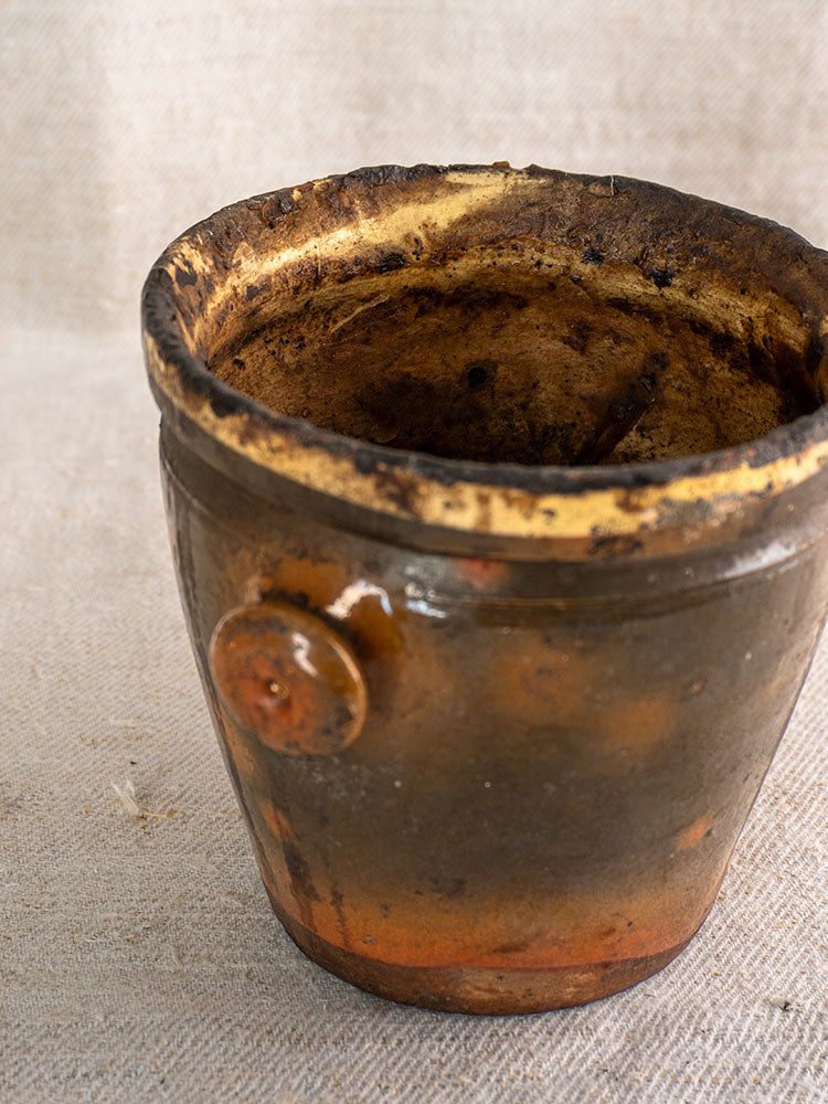 Antique French glazed terracotta pot à graisse with original tag, worn finish and rough rim on textured beige fabric.