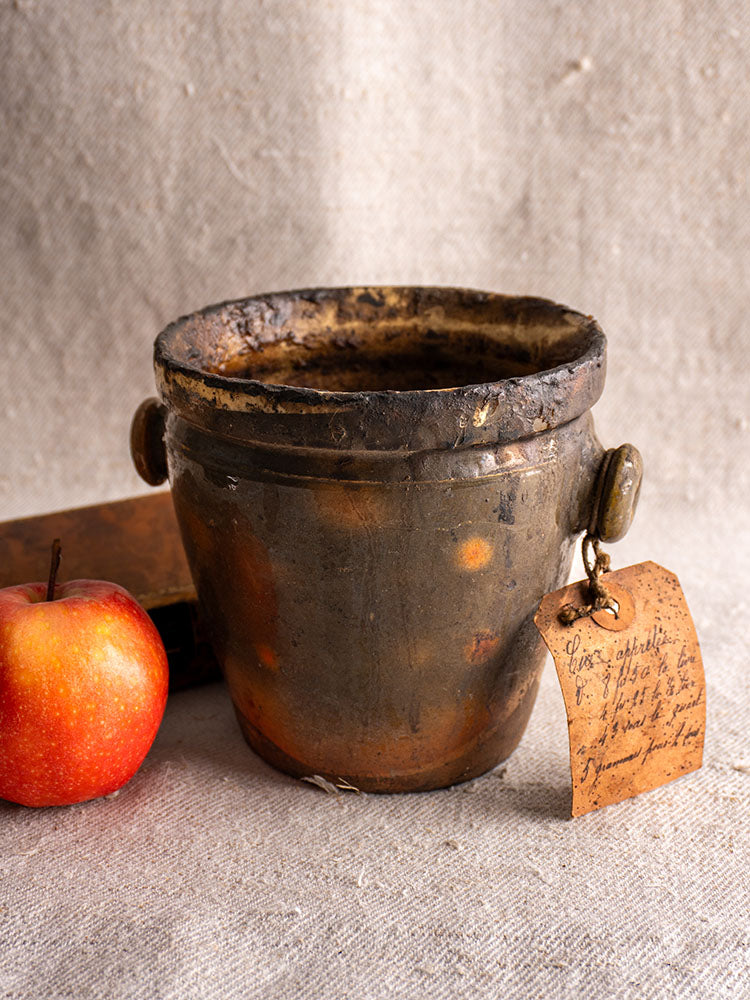 Antique French glazed terracotta pot à graisse with merchant tag and two handles on textured cloth near red apple and closed book.