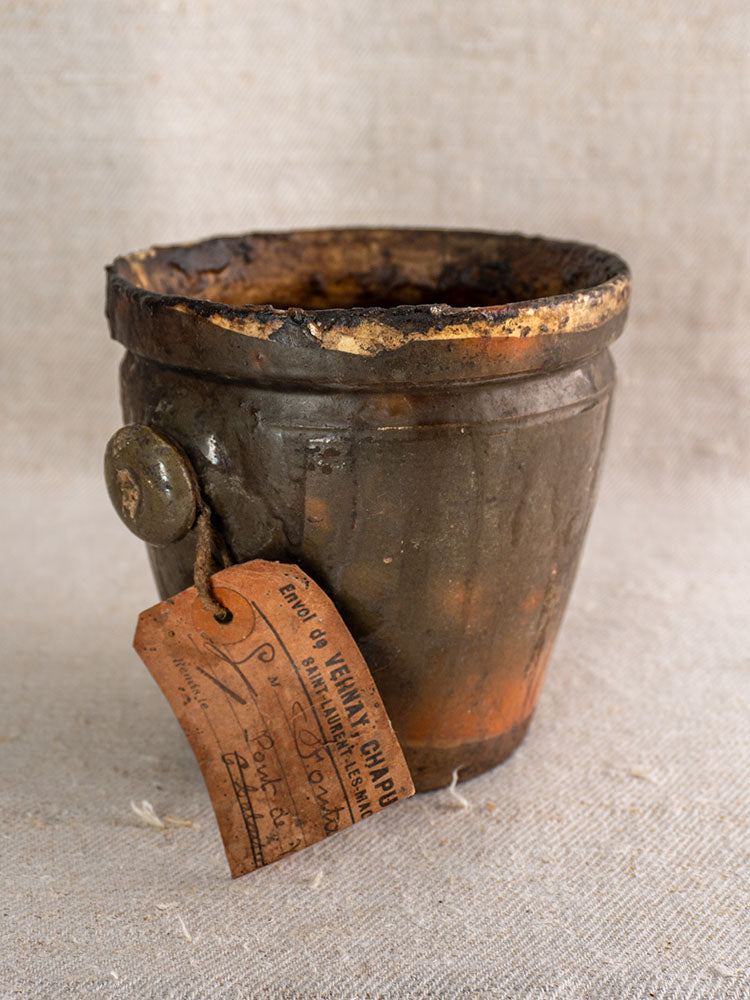 Antique French glazed terracotta pot à graisse with original merchant tag and metal button on textured beige fabric.