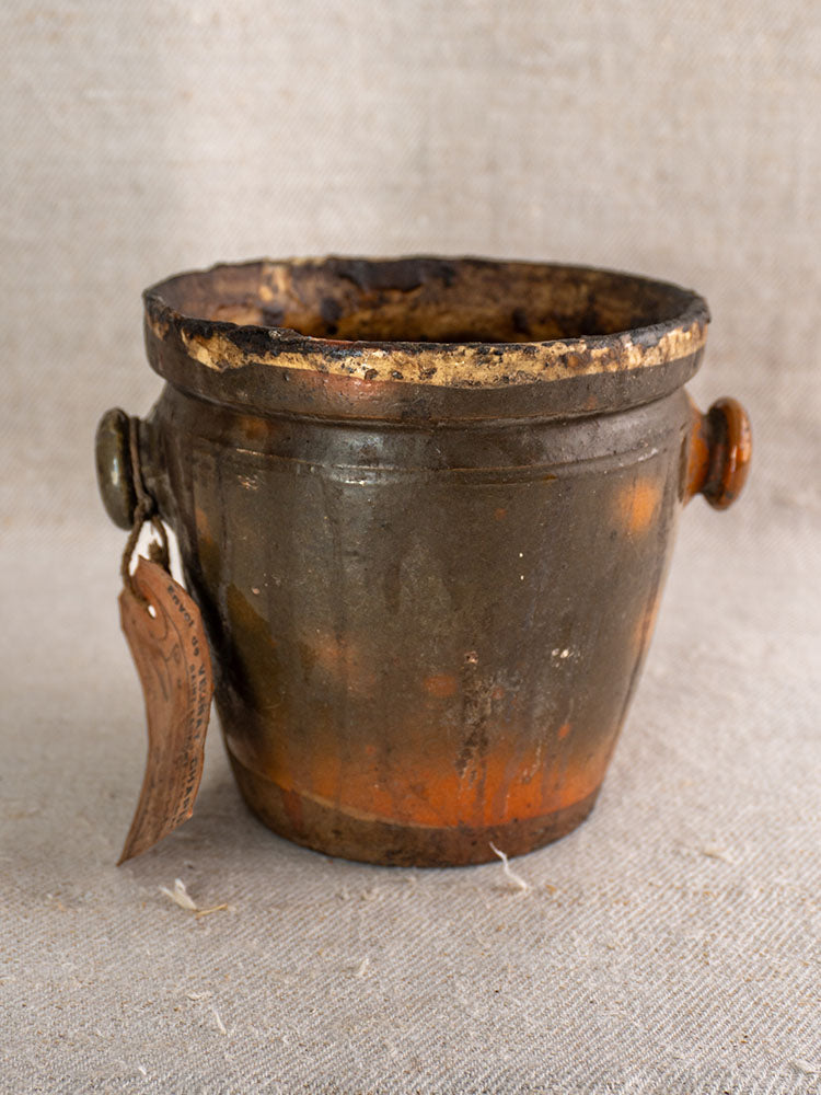 Antique French glazed terracotta pot à graisse with side handles and original merchant tag on beige textured fabric.