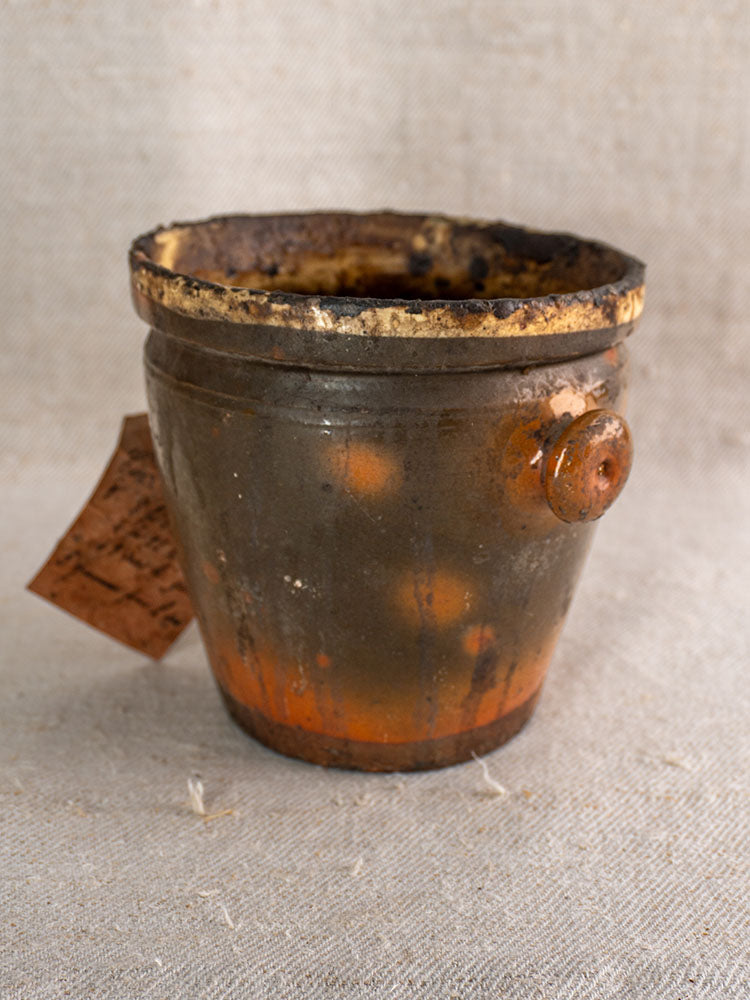 Antique French pot à graisse in glazed terracotta with original merchant tag on textured beige fabric.