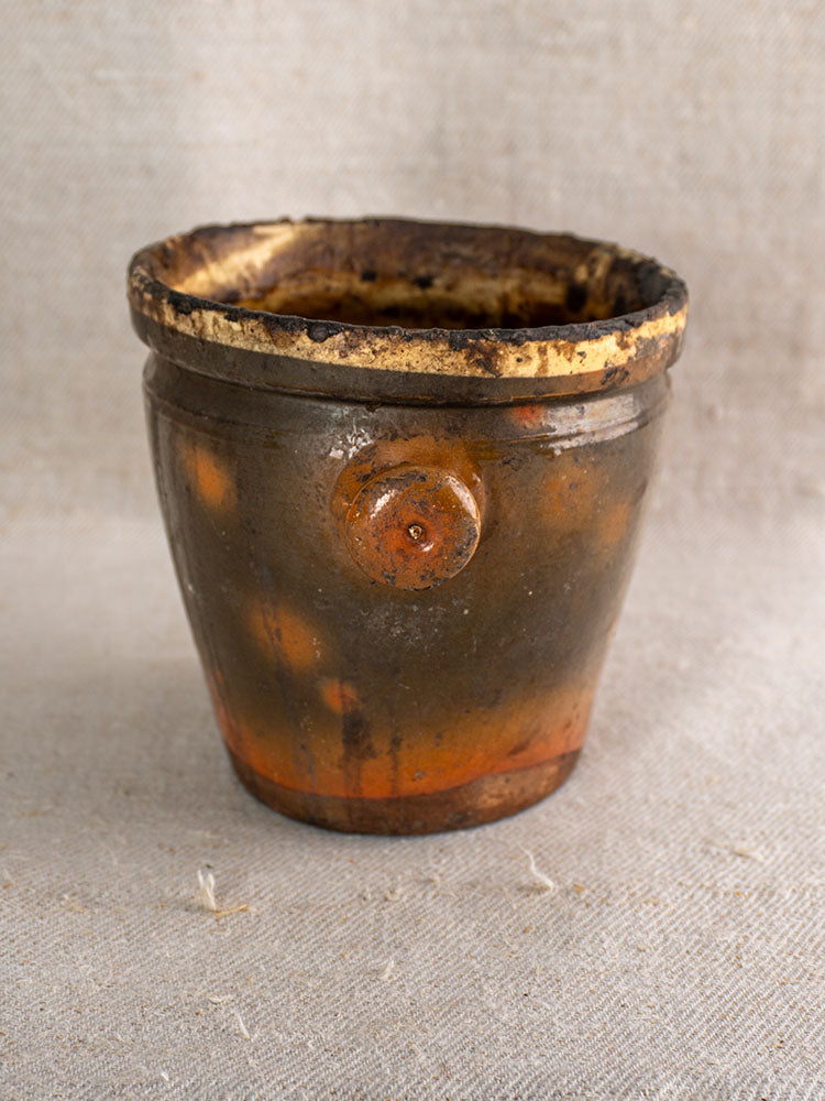Antique French glazed terracotta pot à graisse with merchant tag, earthy brown-orange tones, on textured beige fabric.