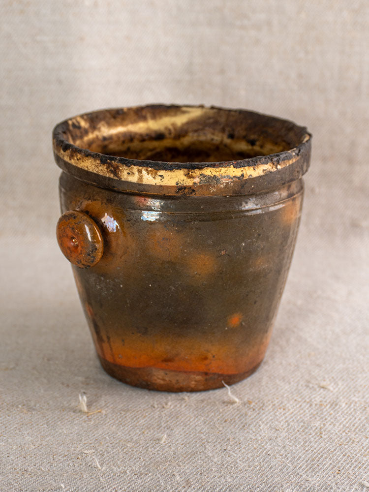 Antique French brown glazed terracotta pot à graisse with weathered rim and merchant tag on beige textured fabric.