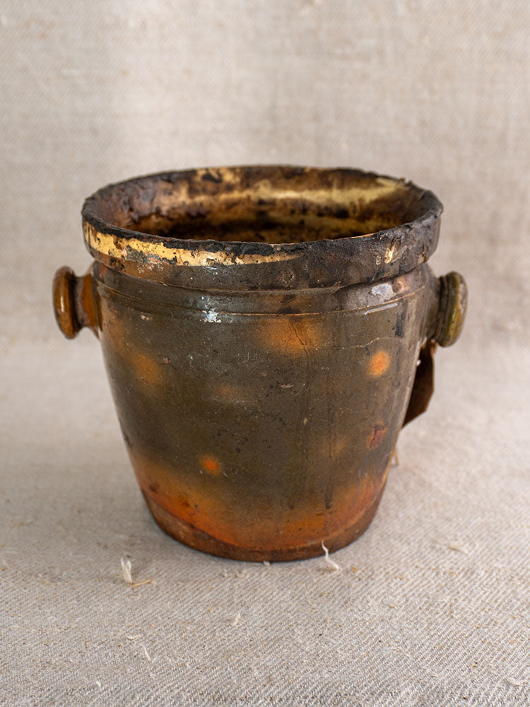 Antique French pot à graisse in glazed terracotta with merchant tag, dark brown and orange, on light textured fabric.