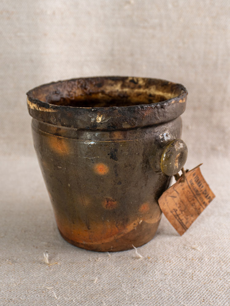 Antique French pot à graisse in glazed terracotta with merchant tag, orange-speckled glaze on textured fabric.