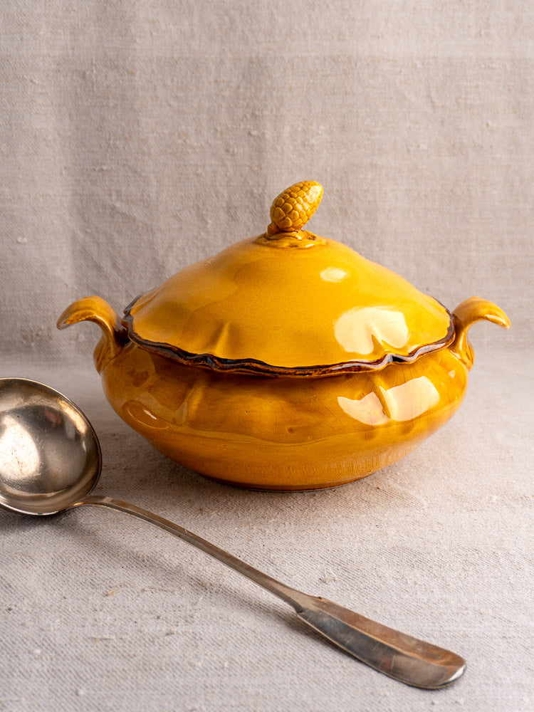 Vintage French Haute Provence yellow faience tureen with pine cone finial and silver ladle on beige fabric.