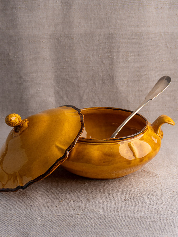Yellow French faience tureen with pine cone finial, lid ajar, metal ladle inside on neutral fabric.