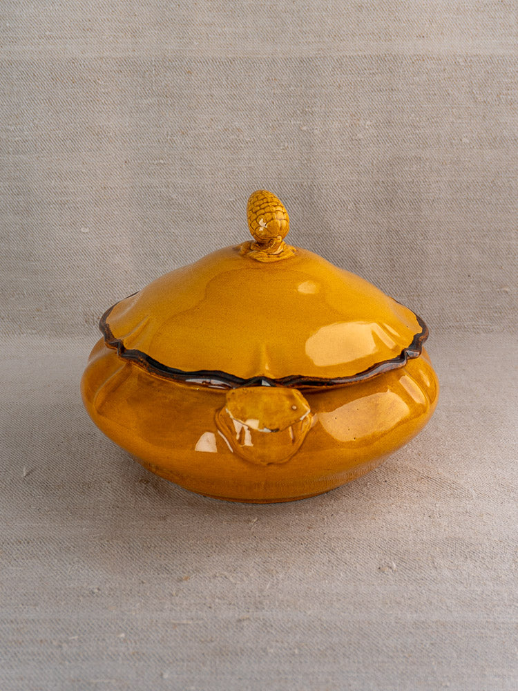 Yellow French faience tureen with pine cone finial and side handle, on neutral fabric background.
