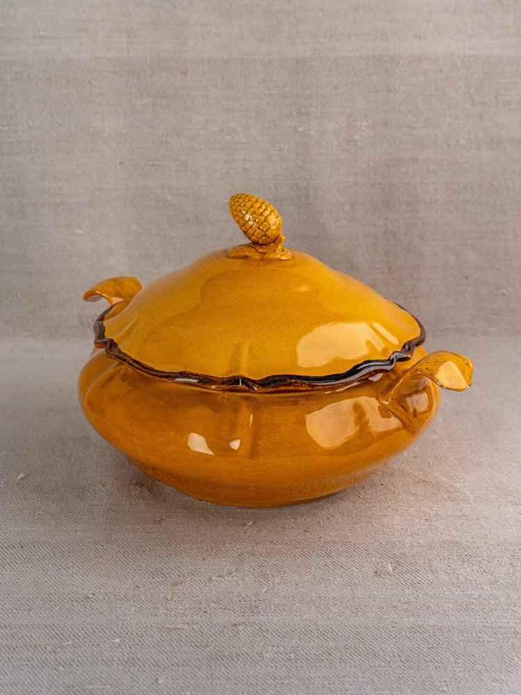 Yellow French faience tureen with pine cone finial on neutral textured fabric background.