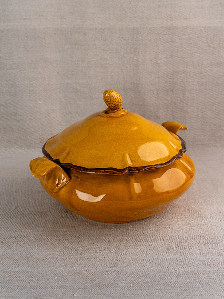 French Haute Provence faience tureen in yellow glaze with leaf handles, pine cone finial, on beige fabric.